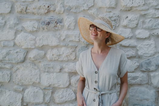 Portrait of beautiful cheerful blonde woman wearing one piece sundress and summer hat, standing in front of old medieval stone wall. Summer vacation portrait concept. Copy space.