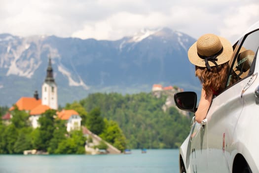 Happy young woman on vacation leaning out of car window on shore of Bled lake, country of Slovenia. Travel, holiday, tourism, explore, rent a car concept