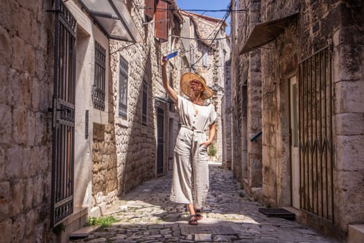 Beautiful blonde young female traveler wearing straw sun hat, taking selfie while sightseeing and enjoying summer vacation in an old traditional costal town at Adriatic cost, Croatia