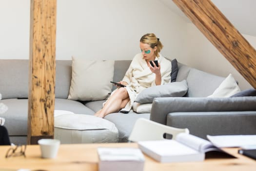 Young female entrepreneur wearing beauty face mask and cosy warm bathrobe in the morning, working remotly from her living room sofa. Remote work from home concept