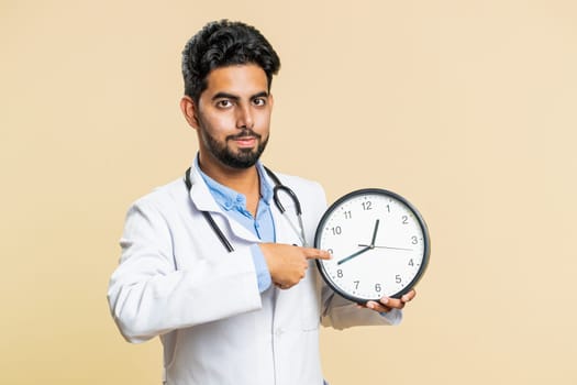 It is your time to heal. Indian young doctor cardiologist man showing time on wall office clock, ok, thumb up, approve, pointing finger at camera. Arabian apothecary pharmacy guy on beige background