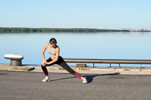 A young woman does a warm-up on the pier in the early morning, against the background of the river. High quality photo
