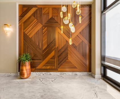 Hall with wood cladding wall decorated with geometrical patterns, tall rounded wooden planter with green bushes, contemporary glass tall chandelier, big window, and white marble floor in modern house