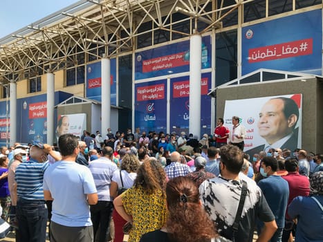 Cairo, Egypt - August 14 2021: Egyptian citizens at Exhibition land waiting for their turn to receive the Covid-19 coronavirus vaccine, with two of the organizers and background of Egyptian president