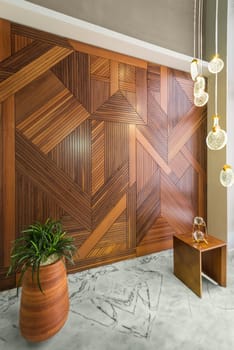 Angled view of hall with wood cladding wall decorated with geometrical patterns, modern glass LED tall chandelier, tall planter with green bushes, and white marble floor in modern house