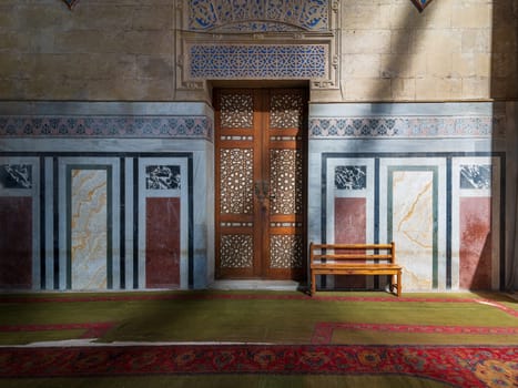 Ornamental wooden door, marble mosaic decorated walls, wooden bench, and floor lit by sunlight beams, located in public historic Al Rifai Mosque, Cairo, Egypt