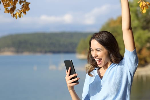 Excited woman checking smart phone celebrating good news outdoors