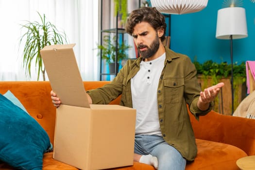 Angry dissatisfied shopper man unpacking parcel feeling upset and confused with wrong mistake delivery from an online store, bad quality broken purchase at home. Arabian guy indoors in room on couch