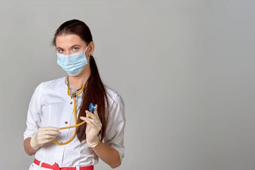 A young doctor a woman in a medical mask in a white coat with a stethoscope in her hands on a gray background, space for text. High quality photo