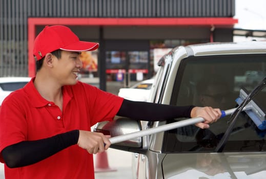 Asian gas station worker in red uniform cleaning the car windshield with window squeegee. A caucasian driver with sunglass waiting inside the car.