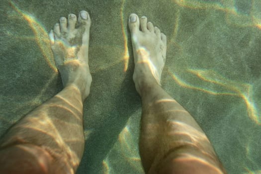 Pair of male feet on sun lit sand sea bottom, view from above, underwater photo.