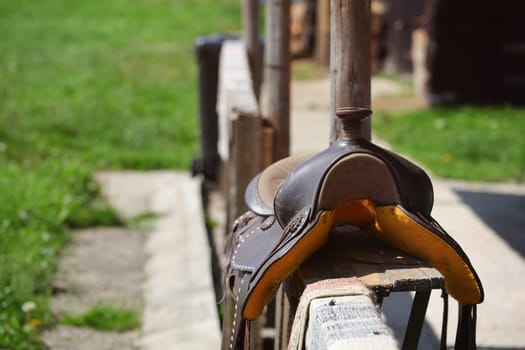 Old worn horse saddle laying on wooden fence lit by sun.