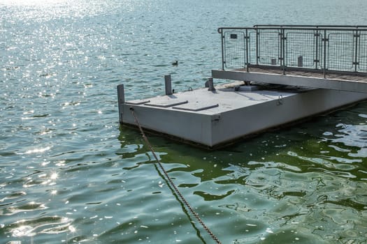 Metal boat pier, on a green water lake, sun sparkling on small waves, chain leading to shore. Brno, Czechia