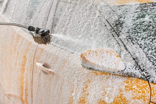 Yellow car being washed in self serve carwash. Shampoo and foam spraying to the side mirror and window.