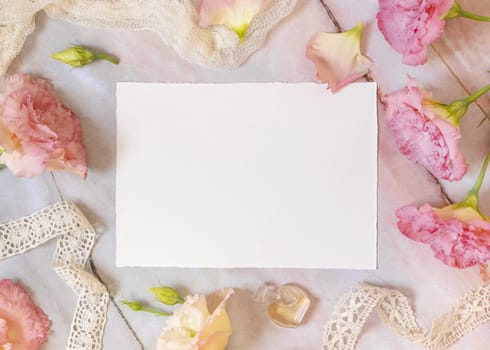 Pink flowers and a blank greeting card laying on a marble table decorated with vintage ribbons, petals and parfume flacon. Mock-up scene. Feminine flat lay