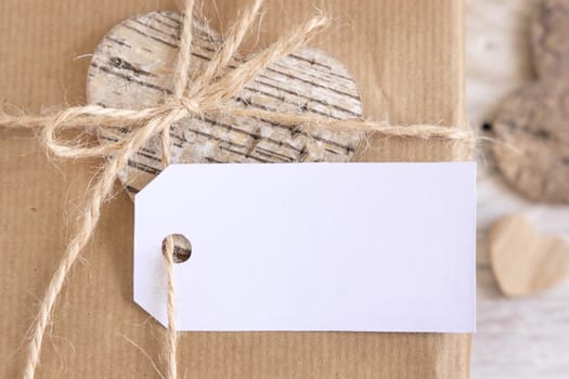 Valentines kraft paper wrapped present with horizontal gift tag hanging on rope bow near hearts top view. Rustic spring scene with blank label mockup, copy space