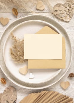 Wedding table place with card and envelope near dried plants, palm leaves, pampas grass and hearts top view. Boho scene with horizontal paper card flat lay. Valentines mockup