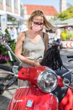 Portrait of casual yound woman wearing protective mask as protection against covid-19 virus using using red retro scooter for her day to day commuting to work