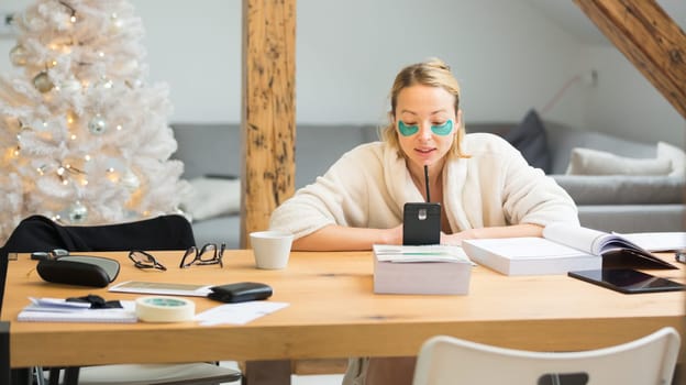 Young businesswoman wearing cosy warm bathrobe having remote cell phone conference call meeting from home living room in winter Christmas time. Remote work from home concept