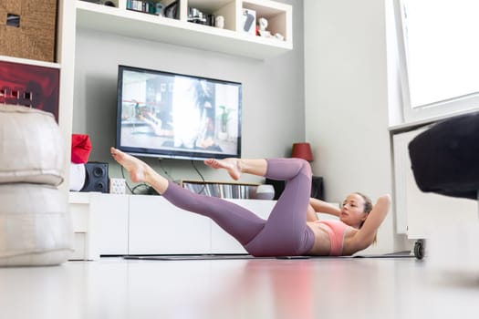 Attractive sporty woman working out at home, doing pilates exercise in front of television in small studio appartment. Social distancing. Stay healthy and stay at home during corona virus pandemic.