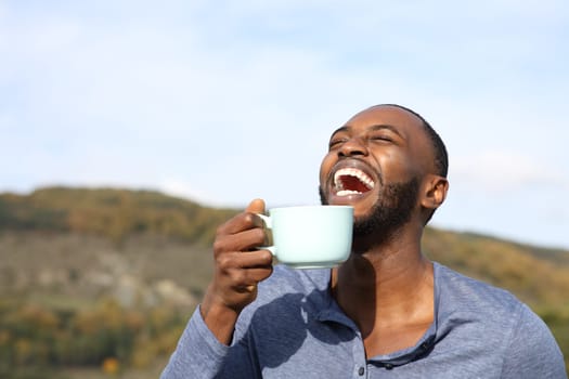 Happy man with black skin laughing and drinking coffee outdoors in the mountain