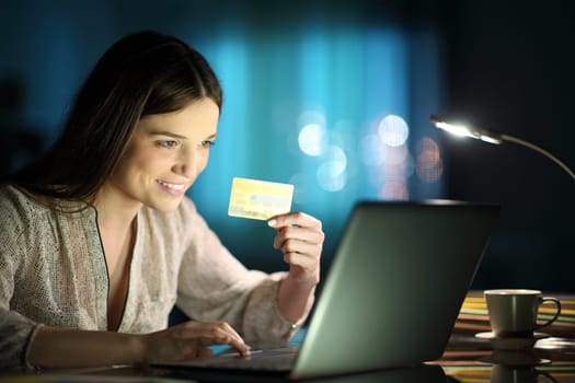 Happy woman holding credit card buying on line with a laptop in the night at home