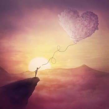A person on the cliff and a heart shaped cloud like a kite raising up in the air. Magical scene, love and romance concept. Pink sunset clouds, peaceful view