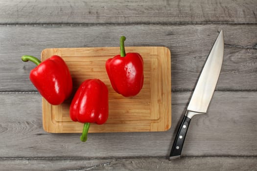 Tabletop view - three whole red bell peppers on chopping board, knife next to it, ready to be cut.