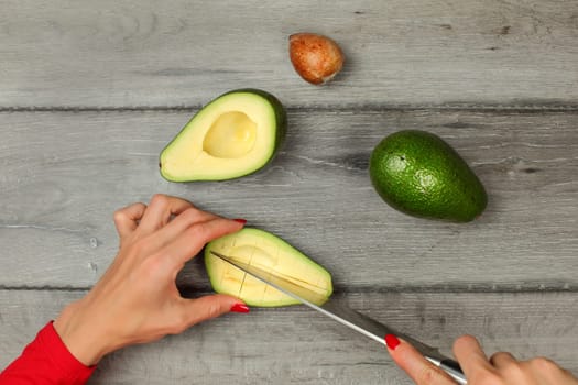 Tabletop view, woman hands holding chef knife and avocado cut in half, seed removed, cutting lines in pulp to portion it.