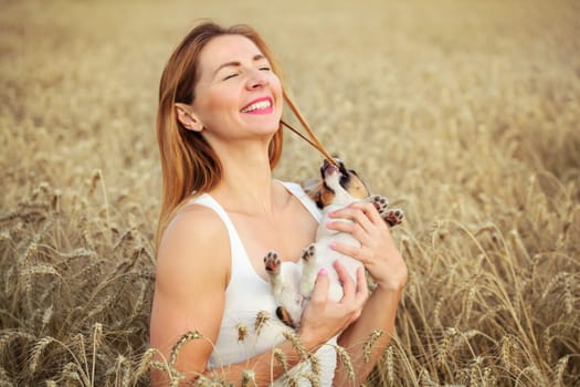 Woman with Jack Russell terrier puppy on her hands, wheat field in background, dog is restless and chewing the hair instead of posing.