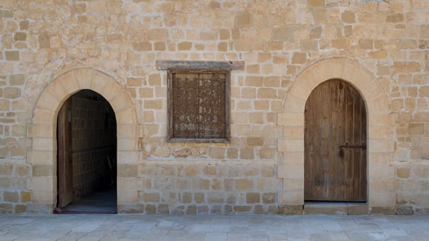 Two weathered wooden arched doors and one closed rusted wrought iron window on bricks stone wall and tiled stone floor