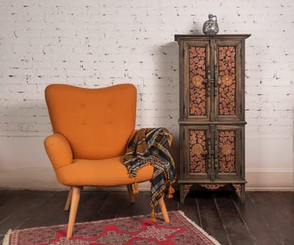 Living room corner with french orange wingback armchair, decorated wooden cupboard, on white bricks wall and grunge wooden parquet with red decorated carpet