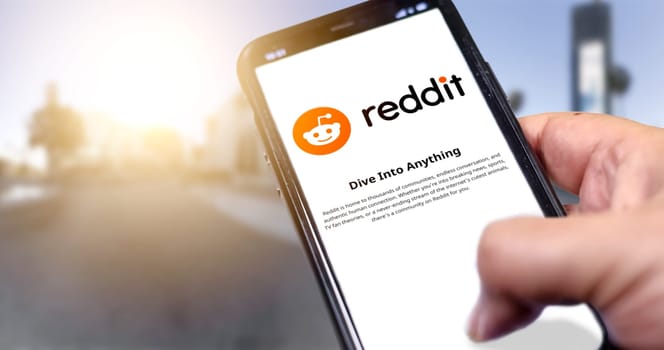Rome, Italy, January 2022: Hand holding a smart phone with Reddit mobile app on the screen on an urban background. Reddit is an American social news aggregation and discussion website application