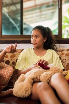 Thai teenager girl sitting on couch with mobile phone and looking to window. Young asian girl looking to side and holding smartphone while sitting on sofa at home. Online education. Using technology. Colourful pillow.