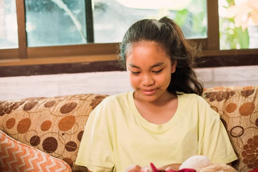 Thai teenager girl sitting on couch with mobile phone and looking to window. Young asian girl looking to side and holding smartphone while sitting on sofa at home. Online education. Using technology. Colourful pillow.