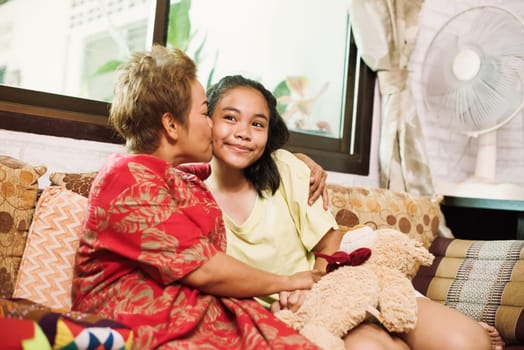 Happy mother kissing and hugging her daughter by sitting on couch at home in Thailand. Beautiful asian family and mothers day concept. Asian family full of love and care. Celebration of graduation