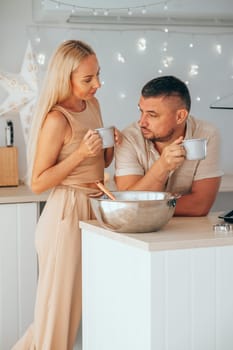 Caucasian couple drinking hot chocolate while preparation dough for Christmas cookies in the decorated kitchen at home. Christmas party preparation. Couple talking while drinking hot cocoa.