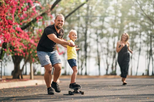 Father holding and training his little son to ride a skateboard and mother is running to them in park on spring time. Concept of leisure and outdor family activity.