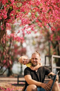 Little boy hugging his sitting father with skateboard in park ander blossom trees. Happy father and son having rest after the skateboarding. Fathers day. Love concept