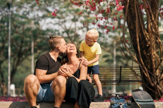 Husband and son is hugging and kissing their wife and mother. Happy family of father, mother and son sitting and hugging each other near skateboard in park. Summer. Family leisure. Love and happiness.
