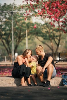 Father and mother is hugging their little son while sitting in skate park with blossom trees around. Concept of family love and care. Outdoor leisure and activity. Happy family.