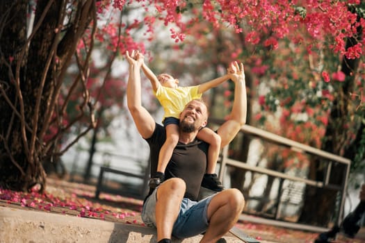 Little boy sitting on the shoulders of his sitting father near skateboard in park and holding hands. Happy father and son having fun after the skateboarding. Fathers day.