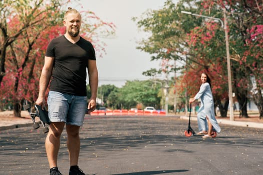Caucasian happy man with trendy hair style holding skateboard and looking to camera in park while his unfocused wife riding scooter behind. Couple enjoying leisure outdoor.