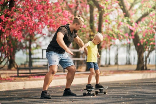 Father holding and training his little son to ride a skateboard in park on spring time. Concept of leisure and outdor family activity. Caucasian man taking care and supporting his kid outdoor.
