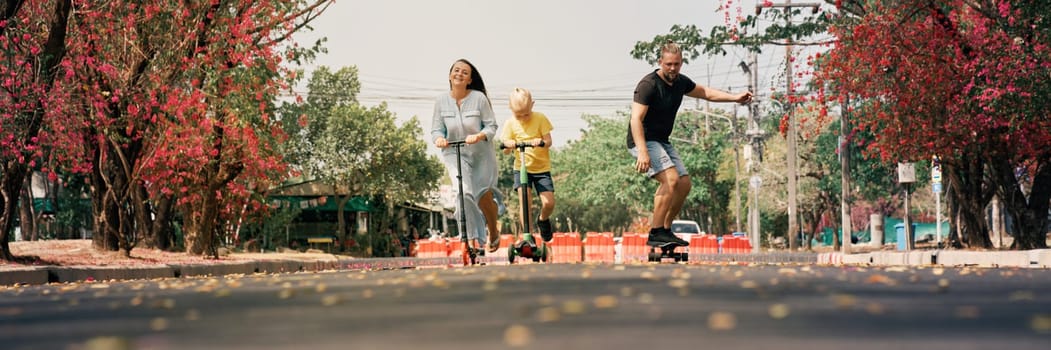 Happy family of father, mother and their little son riding kick scooters with skateboard on pathway of skatepark. Concept of family leisure and outdoor activities.