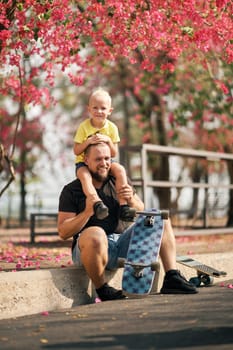 Little boy sitting on the shoulders of his sitting father near skateboard in park. Happy father and son having fun after the skateboarding. Fathers day.