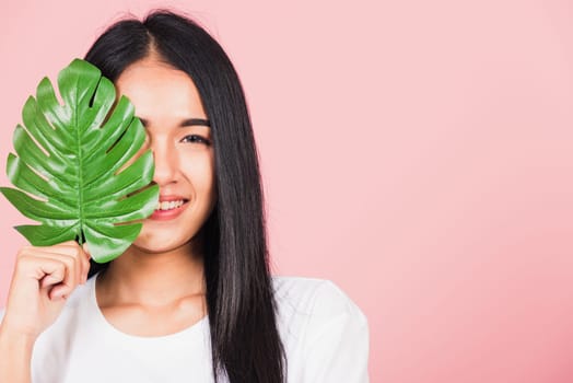 Beauty face. Portrait of Asian beautiful young woman with fresh healthy skin hold green monstera leaf on her face, Tropical Leaves, studio shot isolated on pink background, Skin body care spa concept