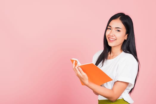 Portrait of happy Asian beautiful young woman confident smiling standing holding orange book open or diary for reading, studio shot isolated on pink background, with copy space, education concept