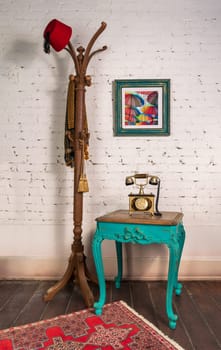 Green wooden vintage side table with golden antique telephone set, and coat hanger stand with red fez and scarf, on room with white bricks wall, hanged painting, and wooden parquet floor