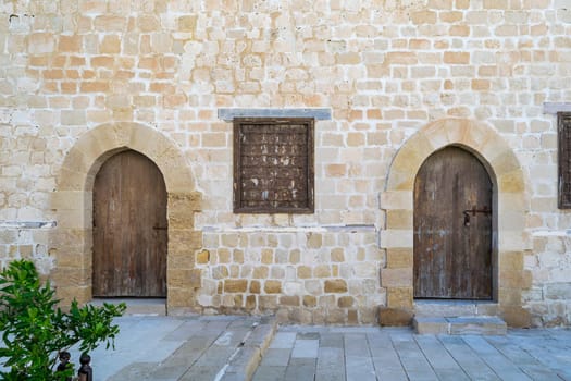 Two weathered wooden arched doors and one closed rusted wrought iron window on bricks stone wall and tiled stone floor